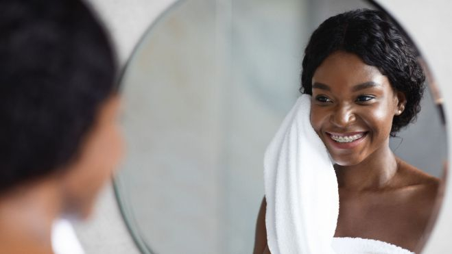 Una mujer sonríe y mira la piel de su cara ante un espejo / Foto: Pexels