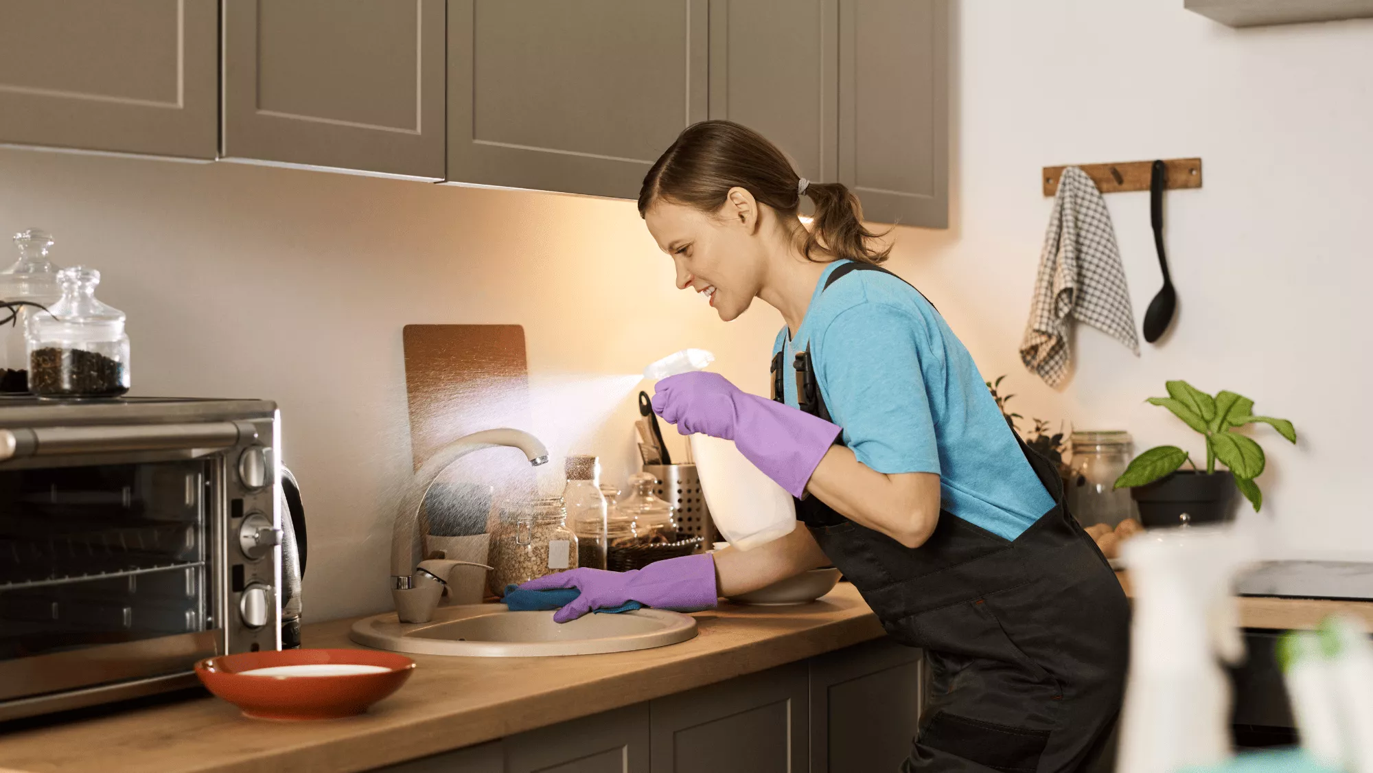 Mujer limpiando el fregadero de su cocina, equipada con guantes y un pulverizador / CANVA